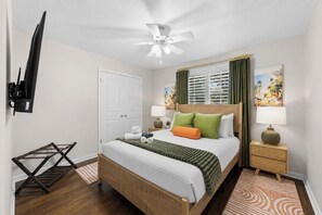 Habitación de hotel con cama, ventilador de techo, televisor montado en la pared y un cuadro en la pared.