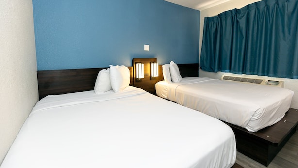 A hotel room with two beds, a headboard with wall-mounted lights, a window with curtains, and an air conditioning unit.