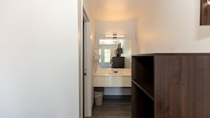 A narrow hallway leading to a bathroom with a sink and mirror, wooden flooring, and a trash bin.