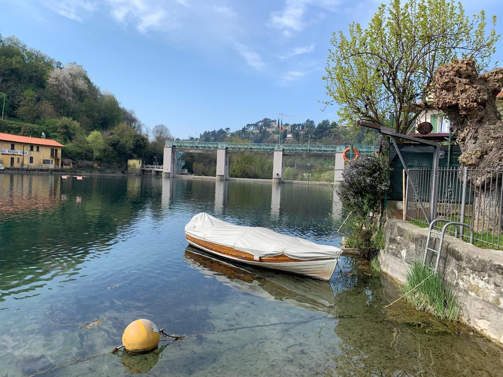 Ein ruhiger Fluss mit einem kleinen Boot, das an der Ufer liegt, einer Brücke im Hintergrund und einem Gebäude mit rotem Dach.