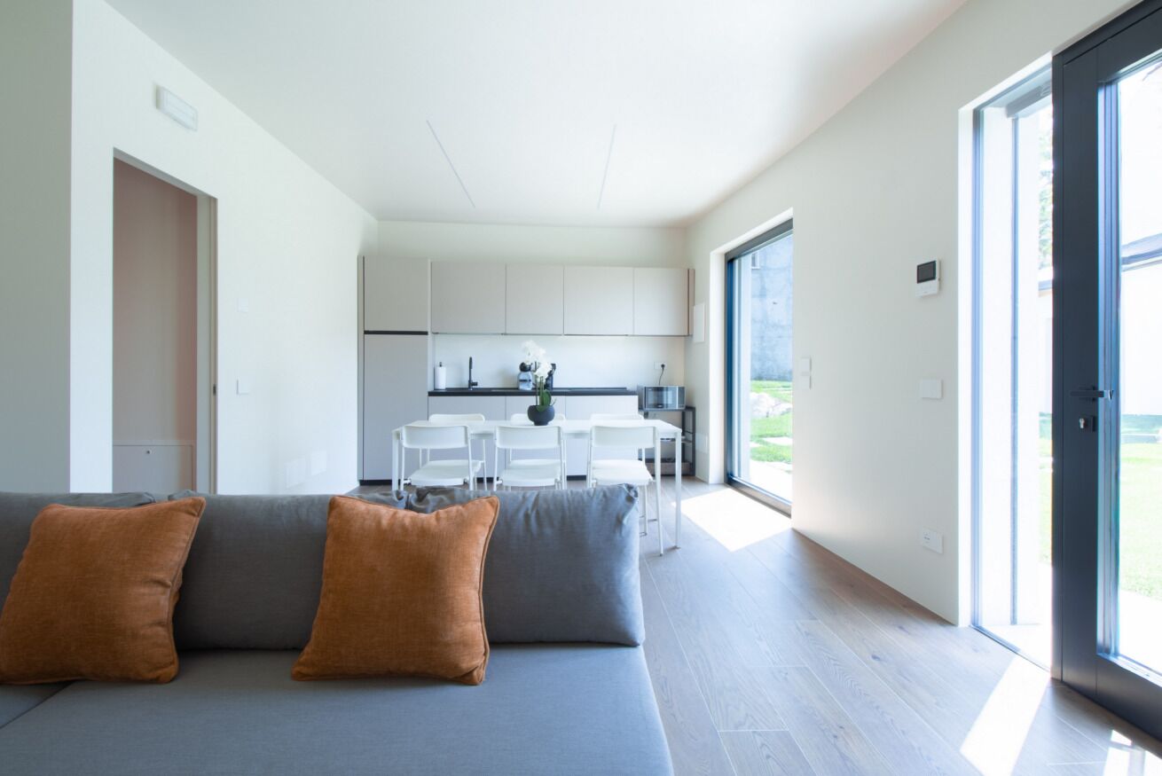 Ein modernes Wohnzimmer mit einem grauen Sofa und zwei braunen Kissen, ein Küchenbereich mit weißen Schränken und einem Esstisch sowie eine große Glastür, die auf einen Balkon führt.