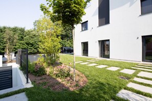 A modern building with a well-maintained lawn, a tree, and a metal fence. - GuestHost - Cozy Apartment with Parking (Venturelli-Fenili)