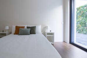 A modern bedroom with a large bed, white nightstands, and a view of greenery through a large window. - GuestHost - Cozy Apartment with Parking (Venturelli-Fenili)