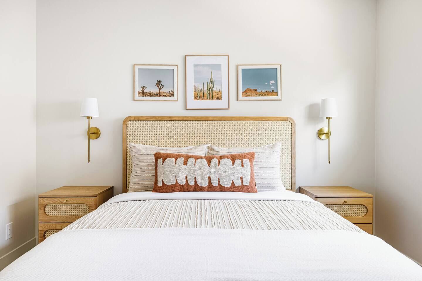 Una cama con una colcha blanca, un solo cojín decorativo y un cabecero con textura tejida. Hay dos mesitas de noche con lámparas, y tres cuadros enmarcados en la pared sobre la cama.