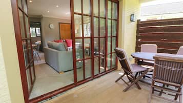 A patio area with a wooden table and chairs, a sliding glass door leading to an indoor living space, and a clock on the wall.
