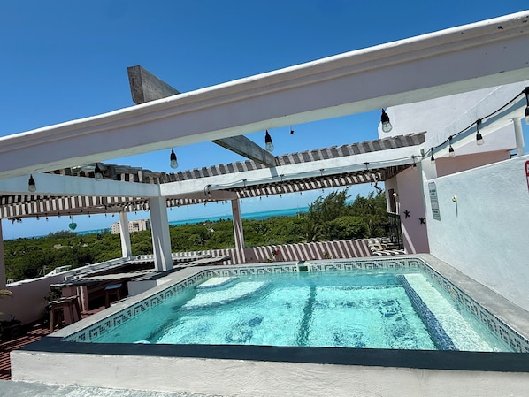 A rooftop swimming pool with a view of the ocean, surrounded by a pergola with hanging lights.