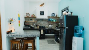 A compact kitchen with a microwave, stove, refrigerator, and a water dispenser. There are wooden stools and a small island with a sink.