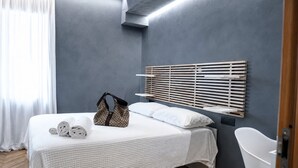 Moderni hotellihuone, jossa on sänky, puinen pääty, valkoinen tuoli ja kylpyhuoneen ovi.