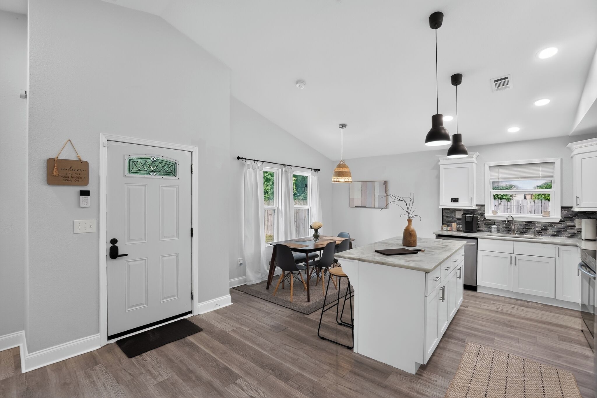 Una cocina moderna con armarios blancos, una isla central, iluminación colgante y un comedor con mesa y sillas.