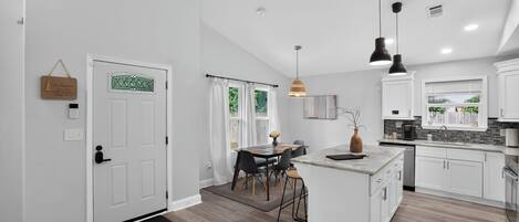 Una cocina moderna con armarios blancos, una isla central, iluminación colgante y un comedor con mesa y sillas.
