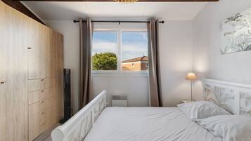 A bedroom with a bed, a wardrobe, a window with curtains, a radiator, and a wall-mounted lamp.