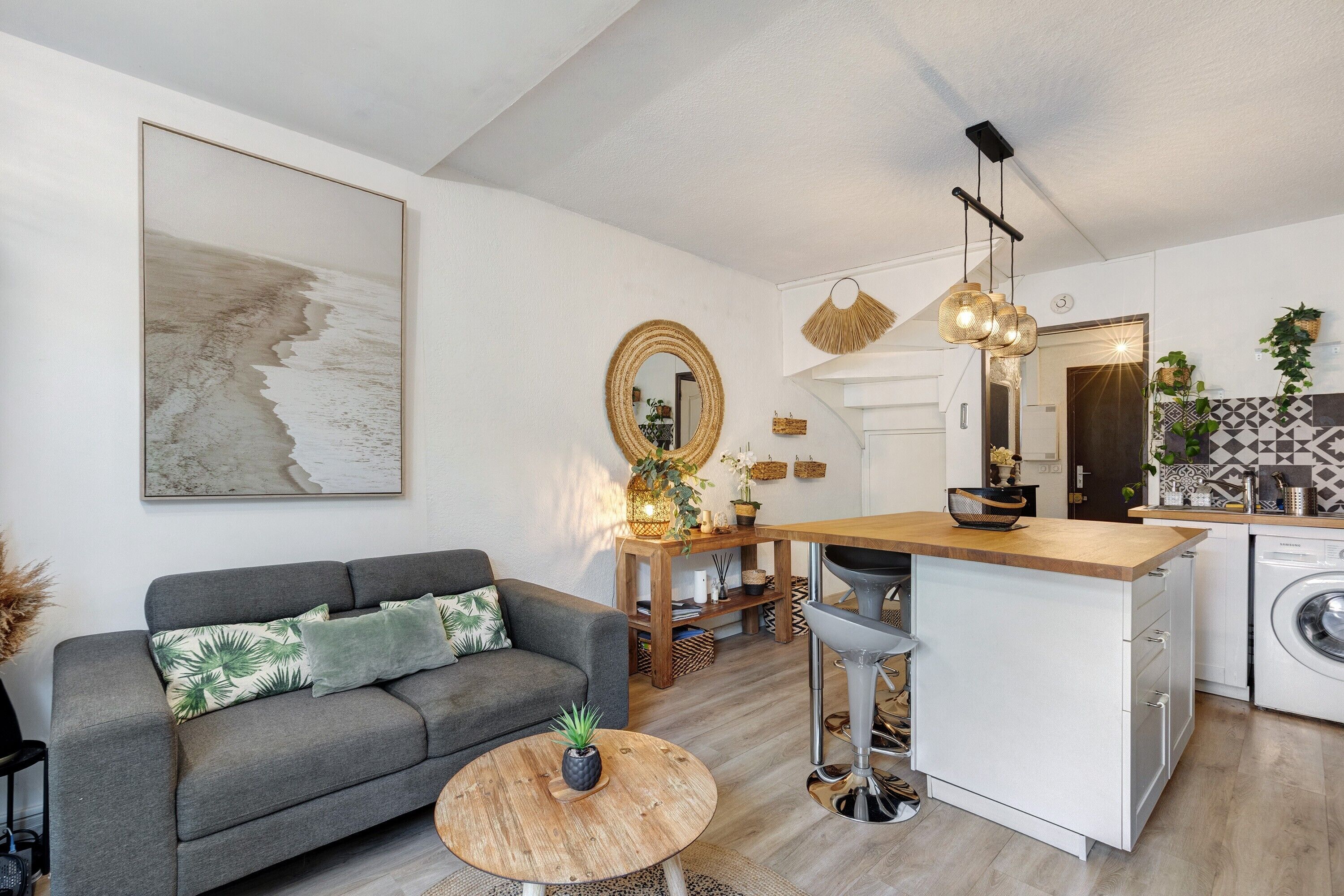 A modern living space with a grey sofa, a round wooden coffee table, a kitchen area with a washing machine, and a dining area with a wooden table.