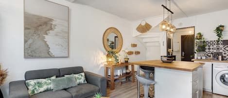 A modern living space with a grey sofa, a round wooden coffee table, a kitchen area with a washing machine, and a dining area with a wooden table.