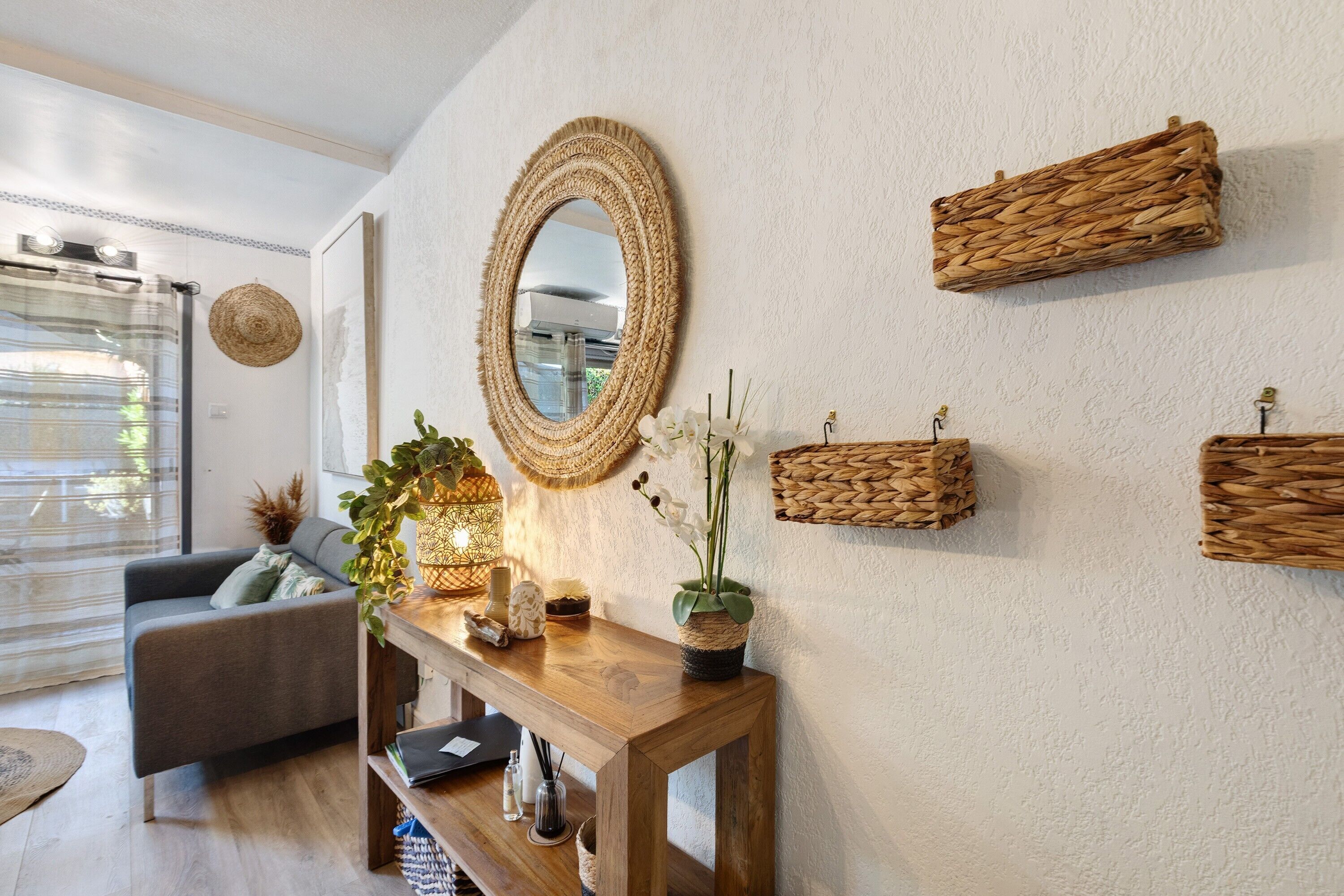 A modern living room with a grey sofa, a wooden console table with decorative items, a large woven mirror, and woven wall hangings.