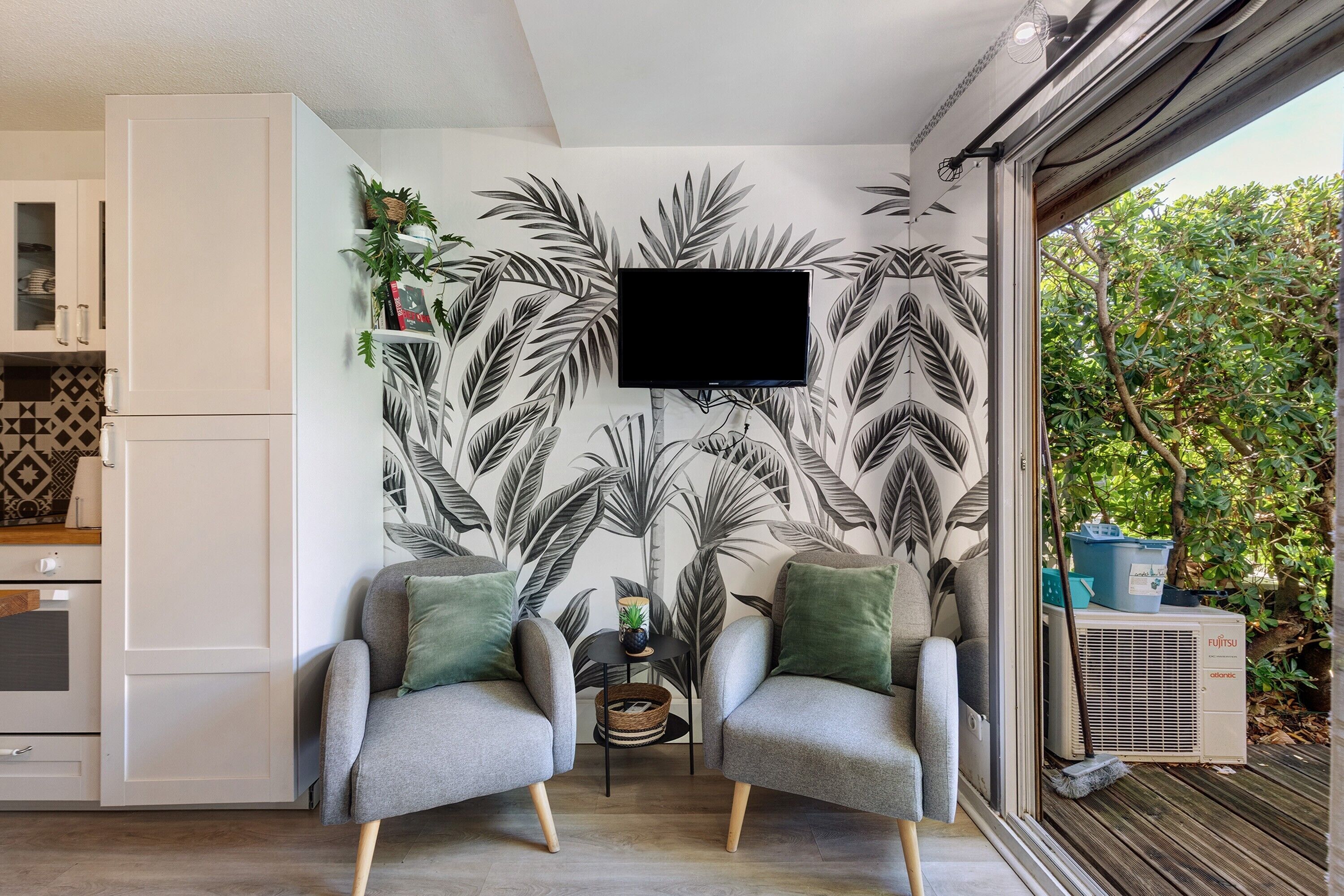 A modern living room with two grey armchairs, a flat-screen TV mounted on the wall, and a large window overlooking greenery.