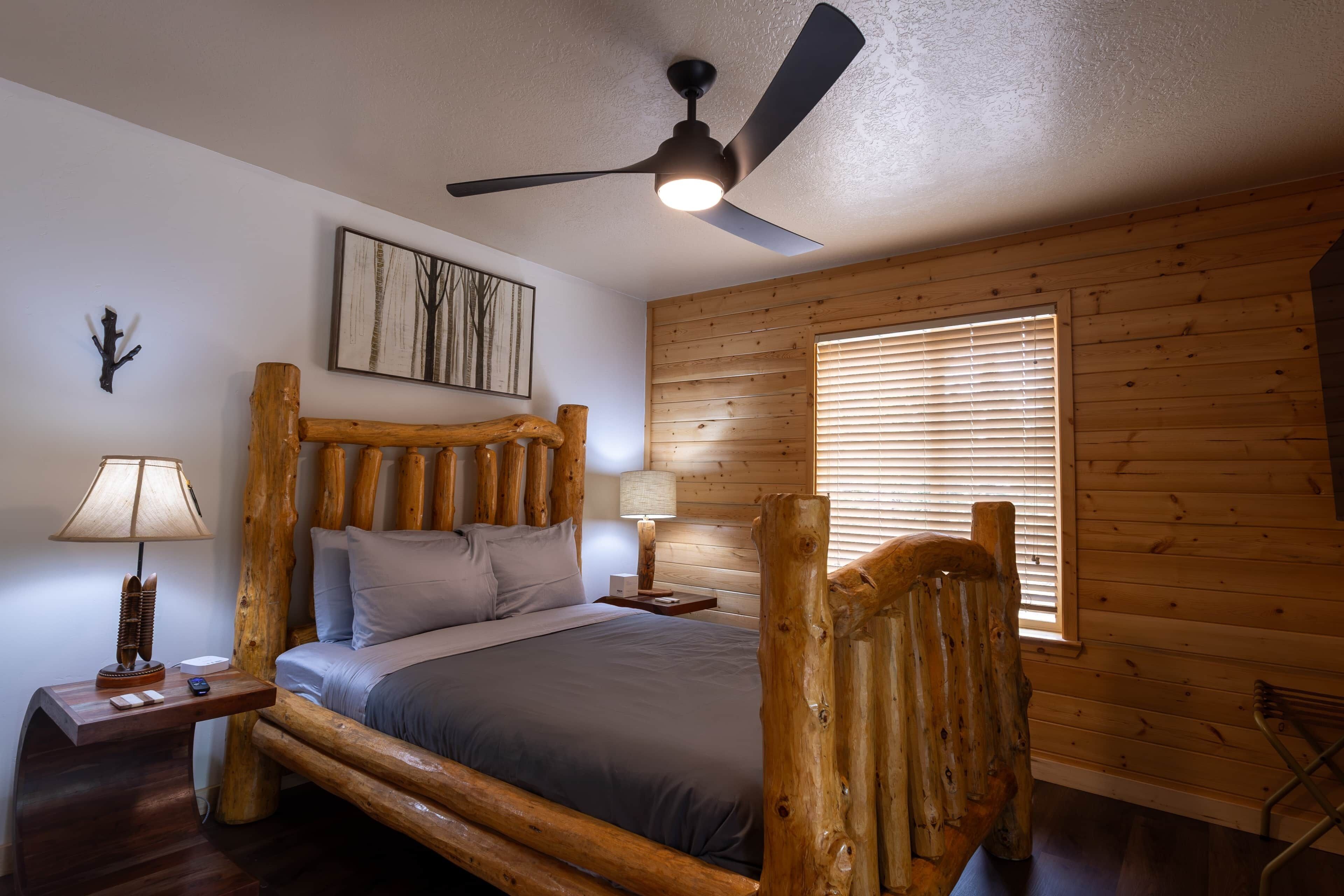 Quarto com estrutura de cama em madeira, ventilador de teto, janela com persianas, mesa de cabeceira com abajur e uma obra de arte pendurada na parede.
