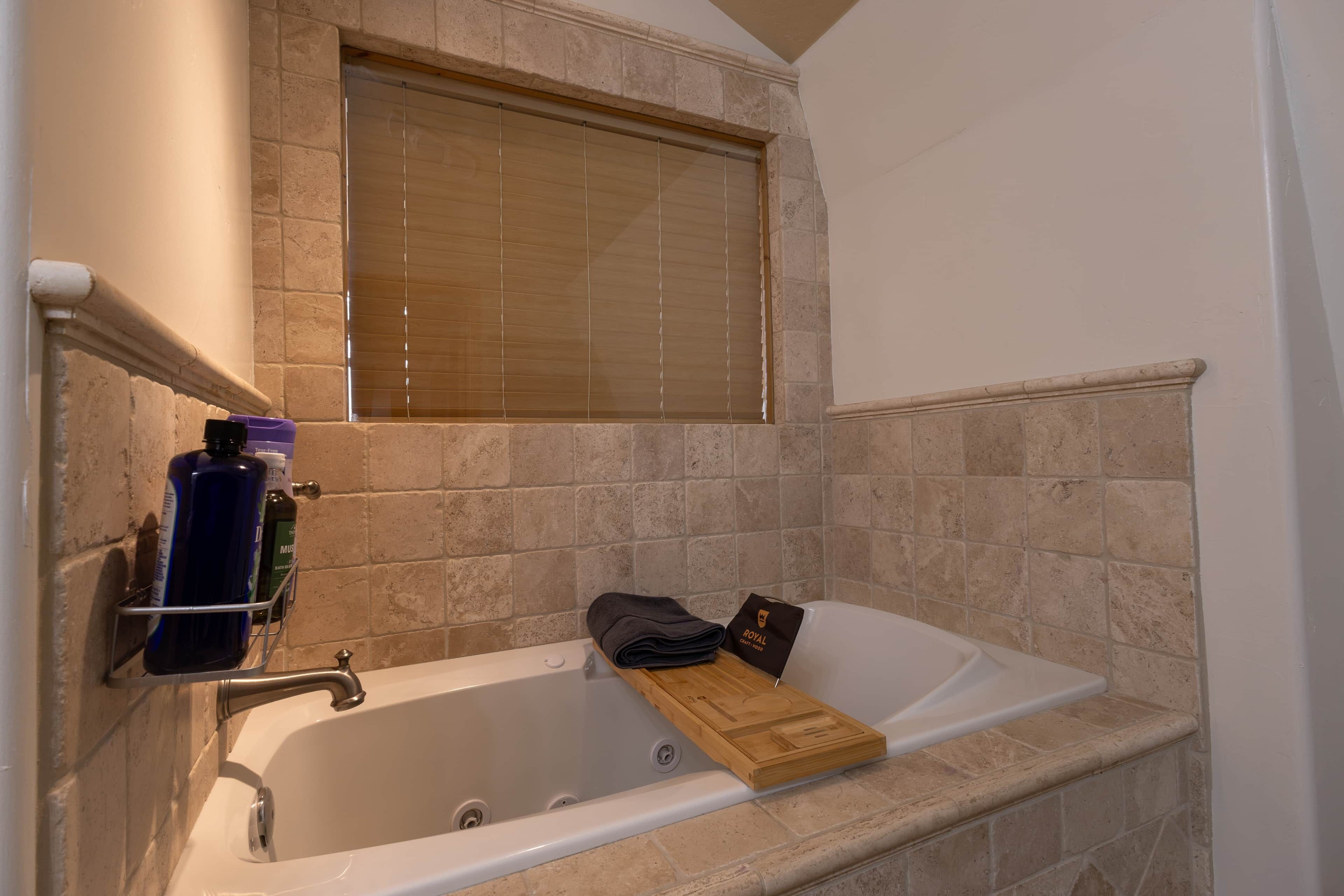 A bathroom with a bathtub, a window with blinds, a towel rack with bottles, and a wooden tray.