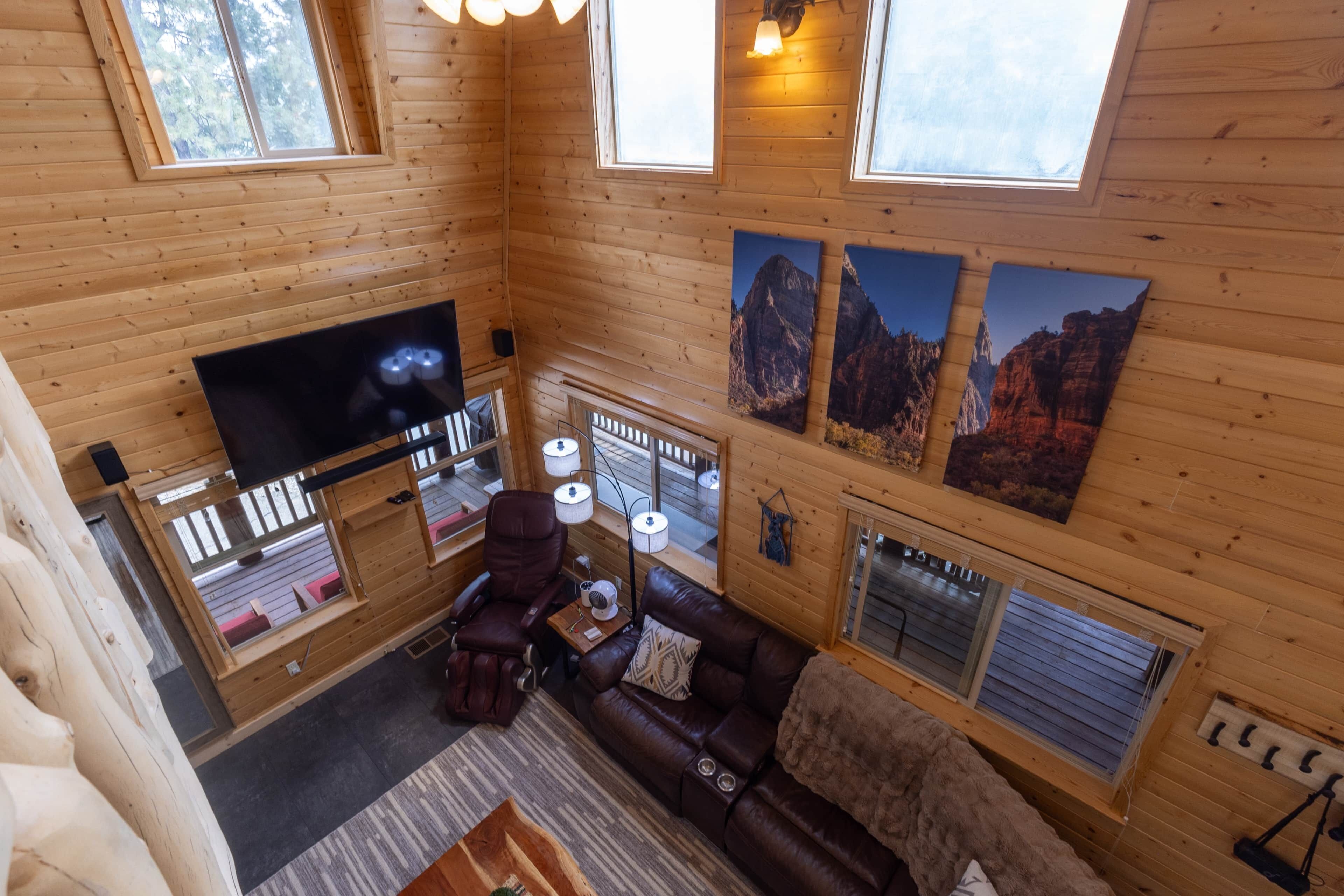 Interior de uma cabana de madeira com televisão de tela plana, sofá de couro e quadros com paisagens de montanha.