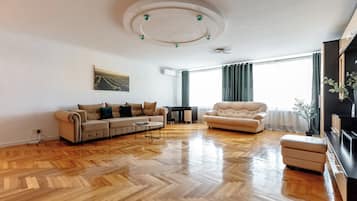 Uma sala de estar ampla com piso de madeira em padrão herringbone, um sofá bege, uma poltrona de couro e uma mesa de centro.