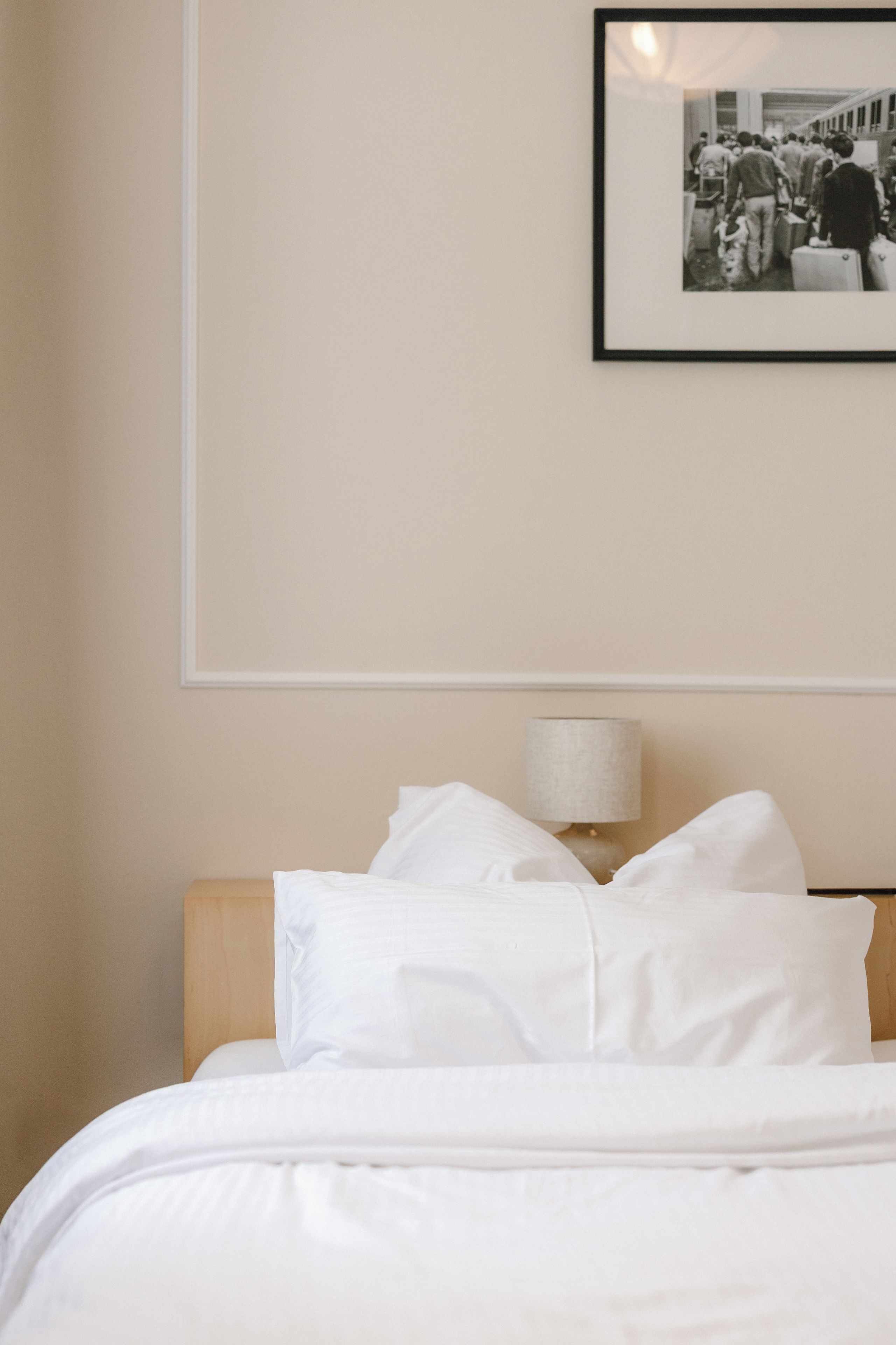 Una cama bien hecha con sábanas y almohadas blancas, una lámpara de noche y una fotografía blanco y negro en la pared.