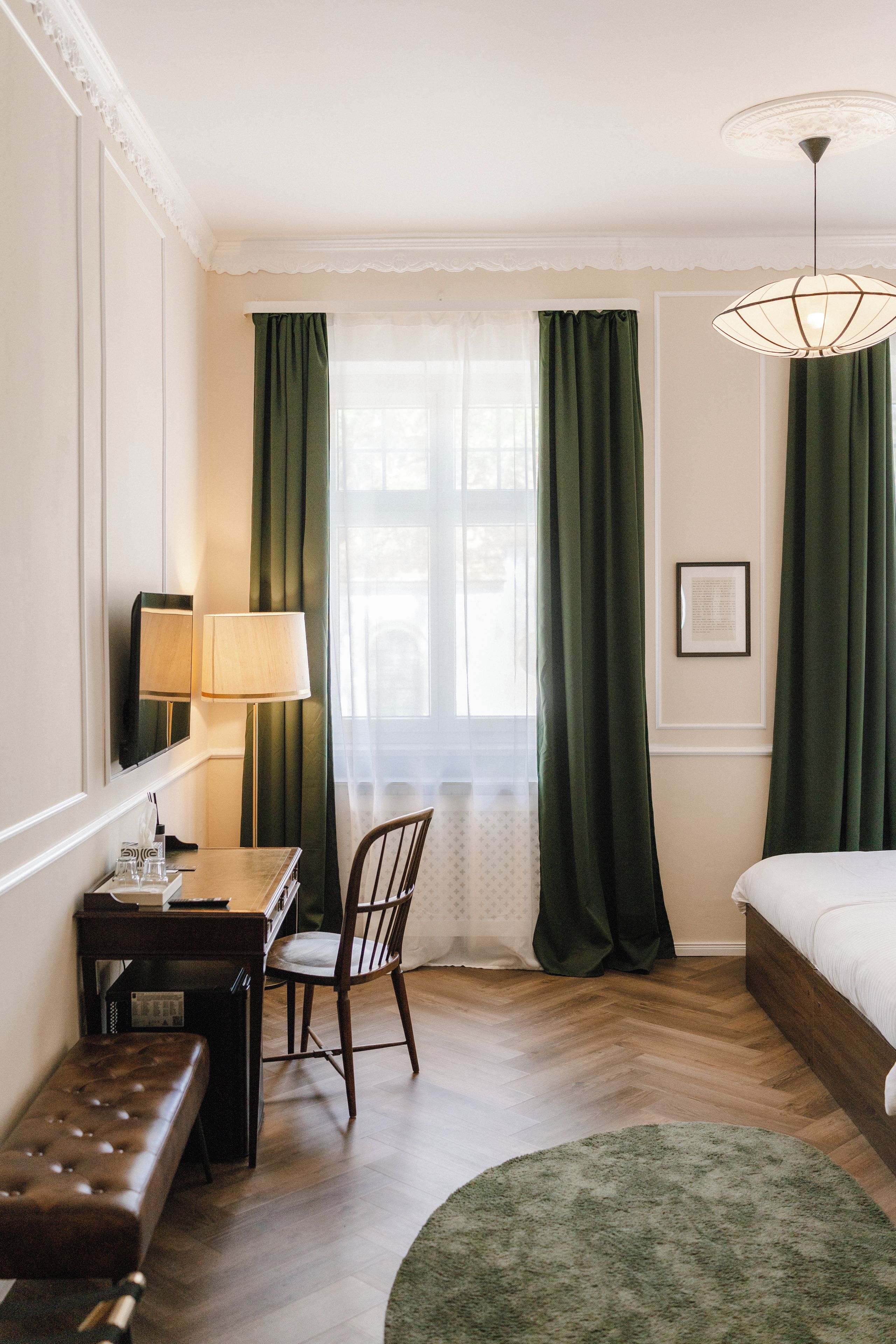 A hotel room with a bed, a desk with a lamp, a chair, a bench, a window with curtains, and a hanging light fixture.