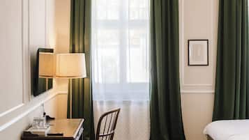A hotel room with a bed, a desk with a lamp, a chair, a bench, a window with curtains, and a hanging light fixture.