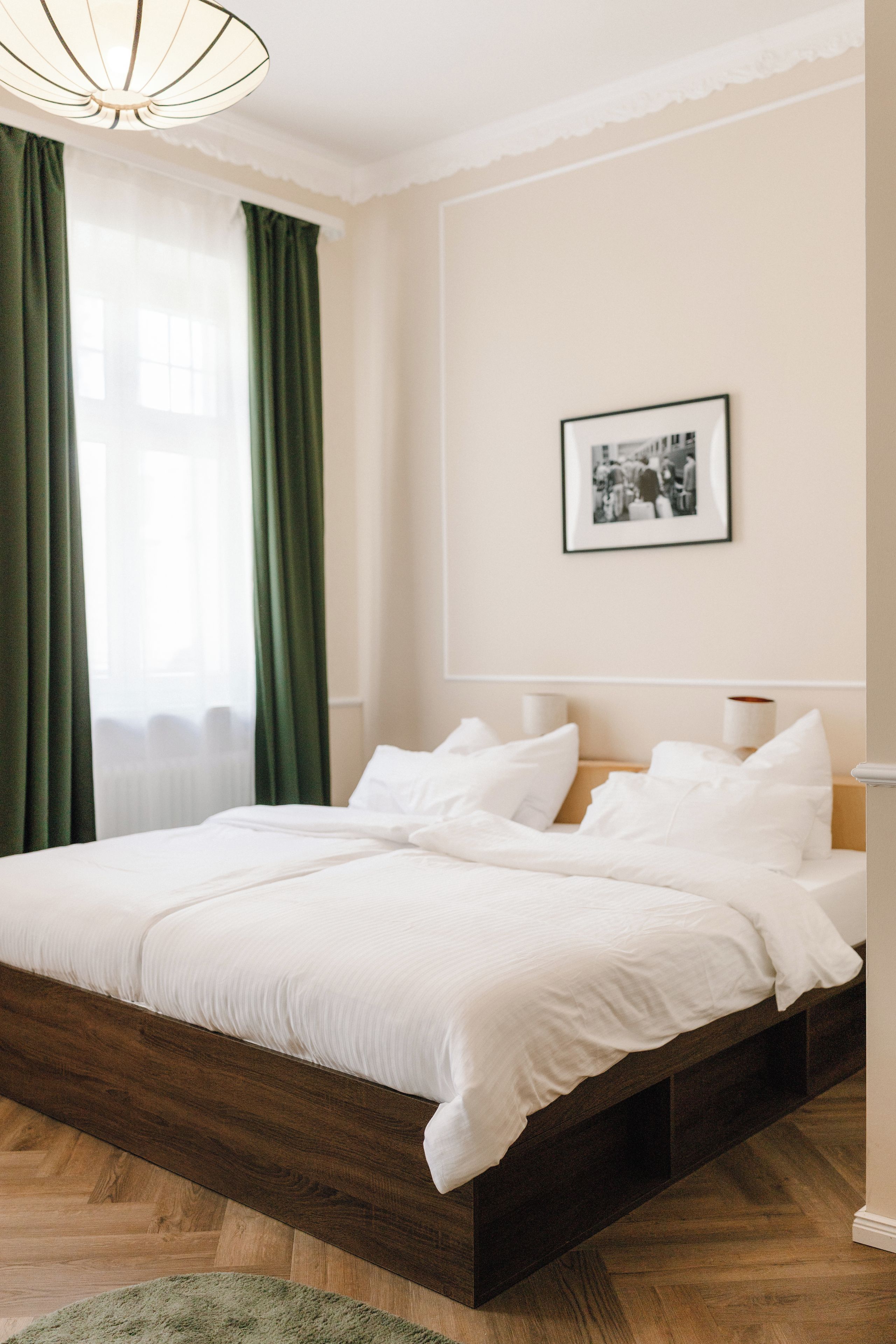 Una cama bien hecha con sábanas blancas en una habitación con piso de madera, una cortina verde y un cuadro enmarcado en la pared.
