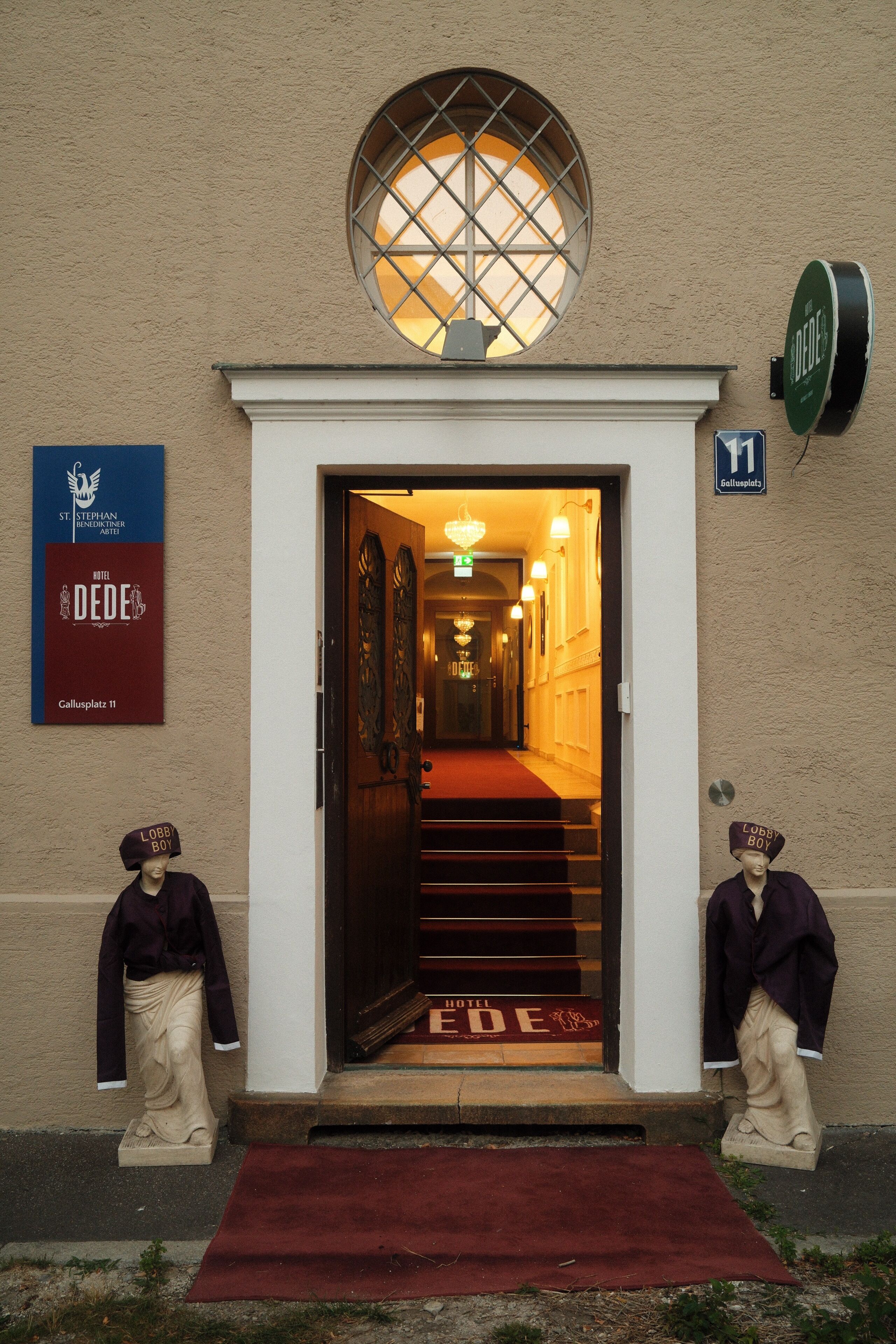 Entrada de un hotel con alfombra roja, una escalera que conduce al interior y dos estatuas a cada lado.