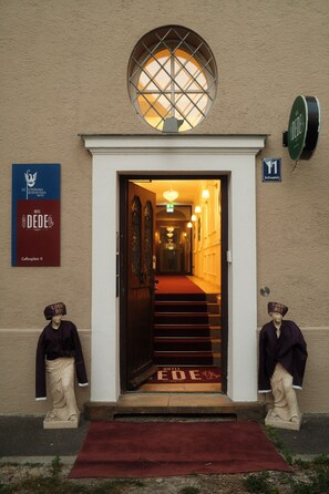 A hotel entrance with a red carpet, a staircase leading inside, and two statues on either side. - Hotel DEDE (Augsburg)