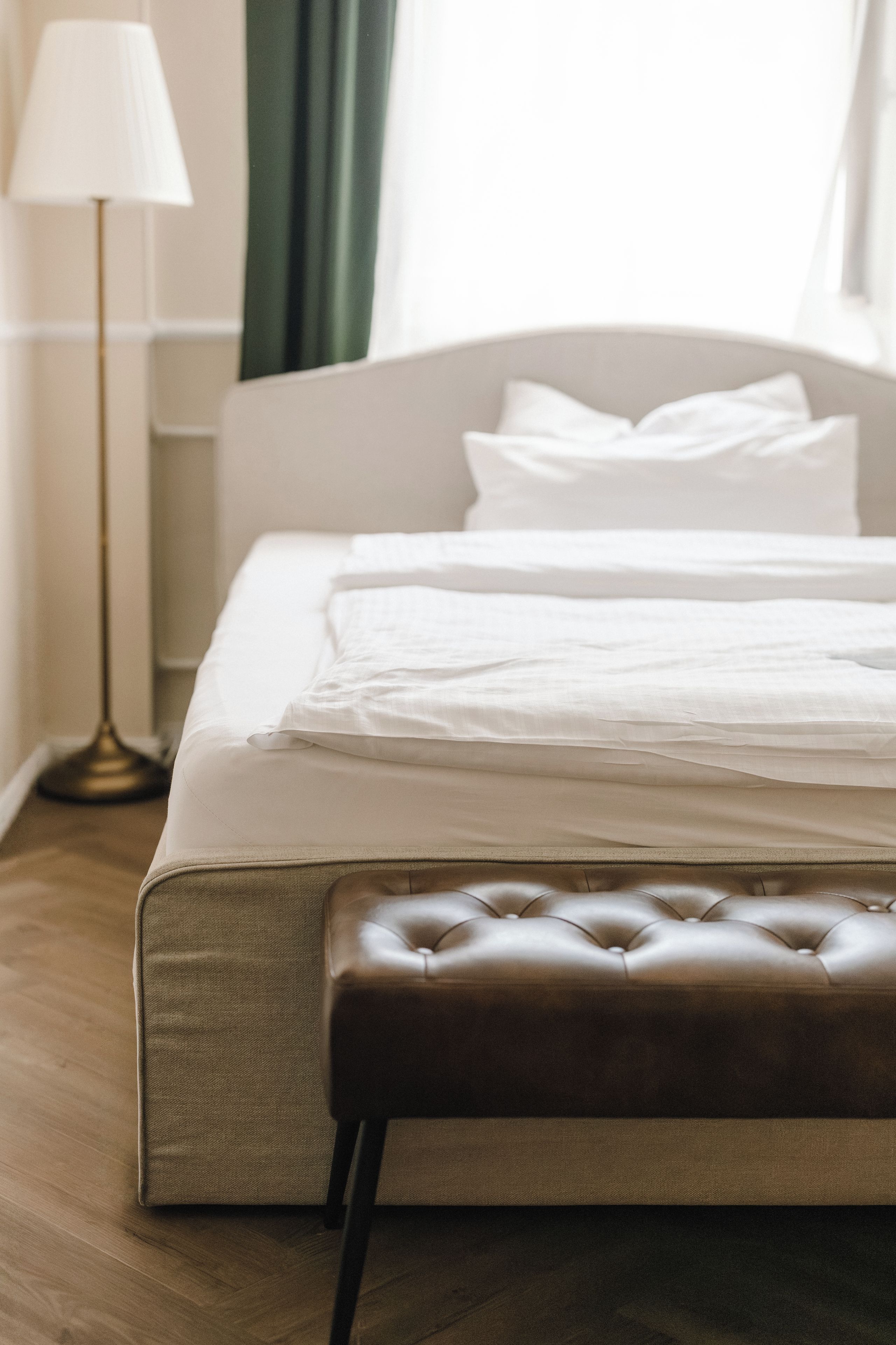 Una cama bien hecha con ropa blanca, un banco de cuero marrón y una lámpara de pie en una habitación de hotel.