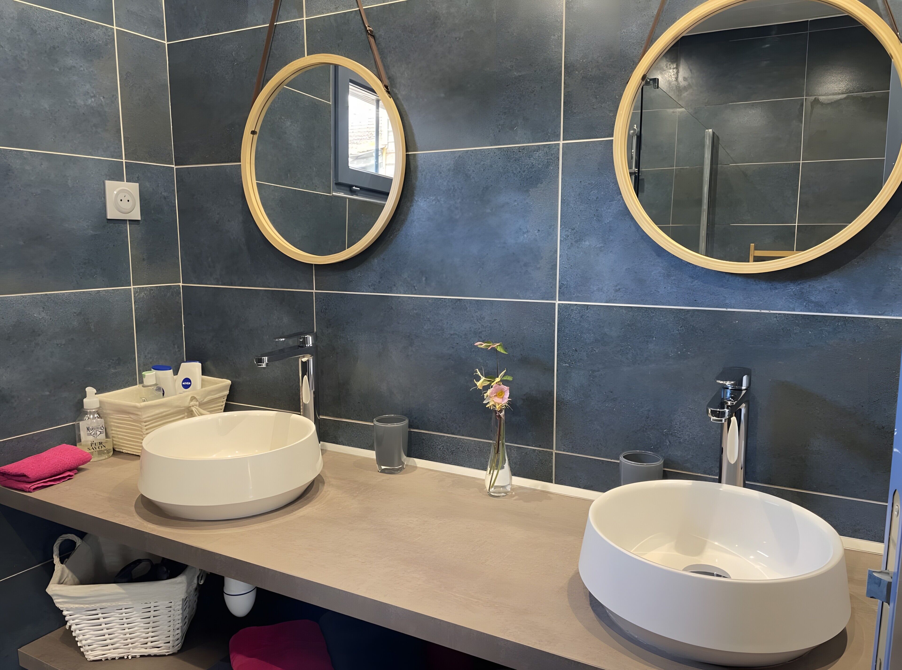 Un baño con dos lavamanos, un espejo y un pequeño florero con flores.