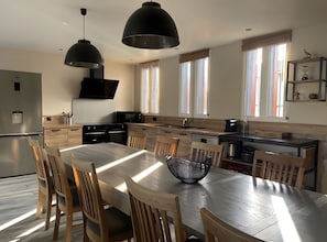 A modern kitchen with a dining area, featuring a large wooden table, chairs, pendant lighting, and a built-in oven.