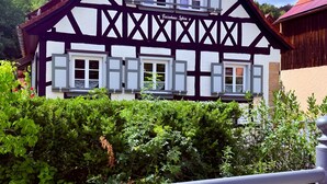 Een klassiek vakwerkhuis met veel ramen en een balkon, omgeven door weelderige begroeiing.