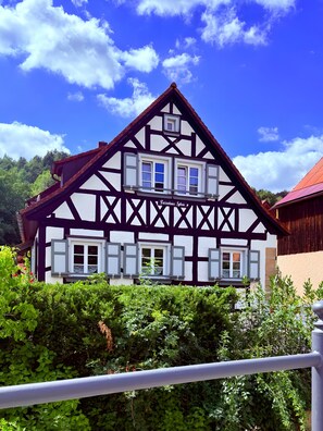A traditional half-timbered house with multiple windows and a balcony, surrounded by lush greenery.
