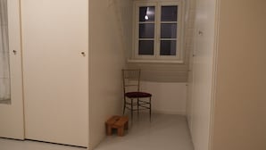Een smalle gang met een houten stoel en een klein houten krukje, een raam met zonwering, en een deur.