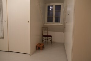A narrow hallway with a wooden chair and a small wooden stool, a window with blinds, and a door.