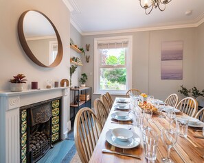 Un comedor con una larga mesa de madera dispuesta para una comida, una chimenea con azulejos decorativos y un espejo grande en la pared.