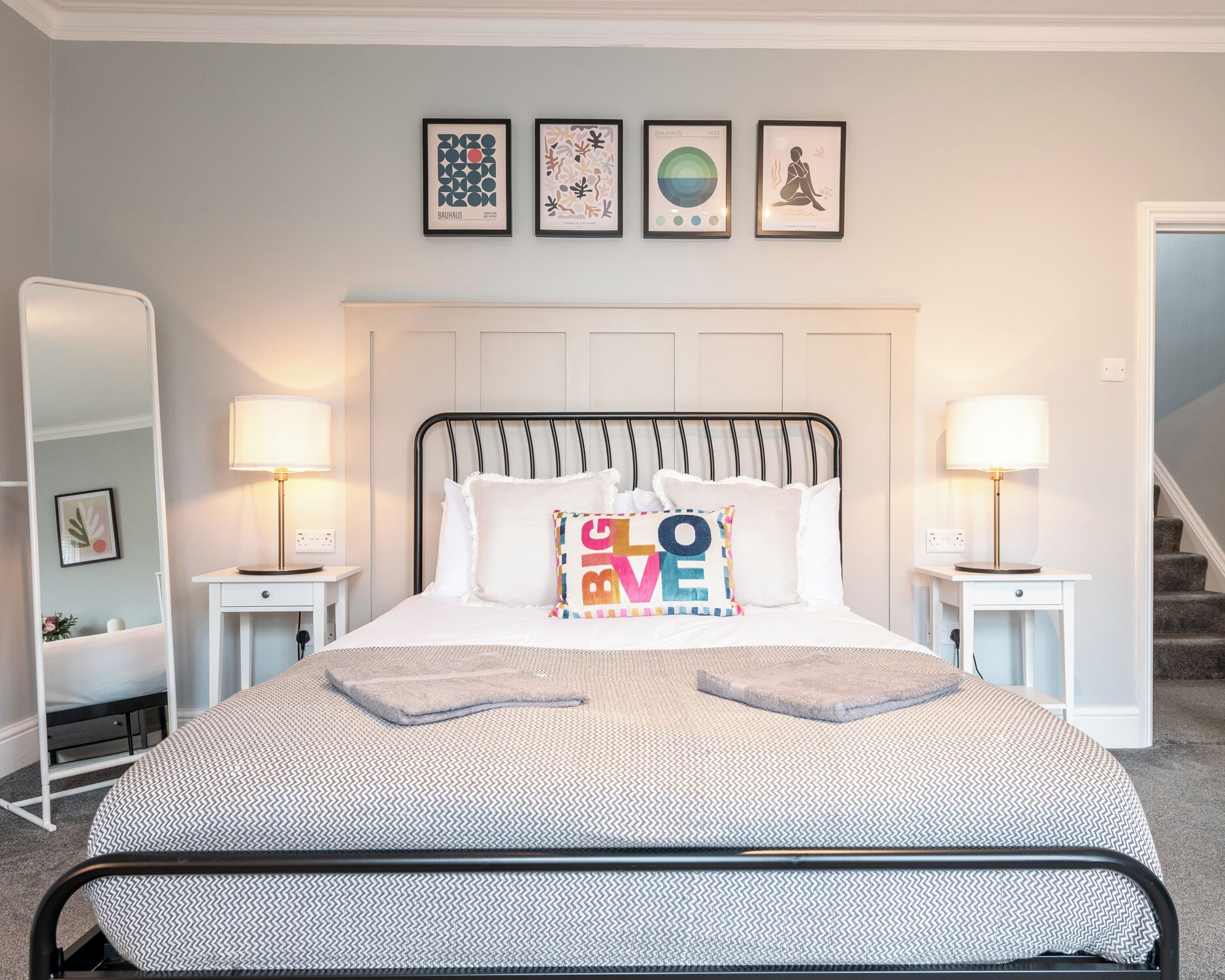 A neatly arranged bedroom with a bed, bedside tables, a mirror, and wall art.