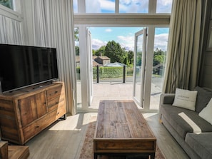 A living room with a television on a wooden cabinet, a sofa with a cushion, and a view through a glass door to the outside. - Dales Retreat (Skipton)