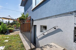 A residential building with a blue exterior, a small window with a decorative grill, a metal railing, and a potted plant with red flowers.