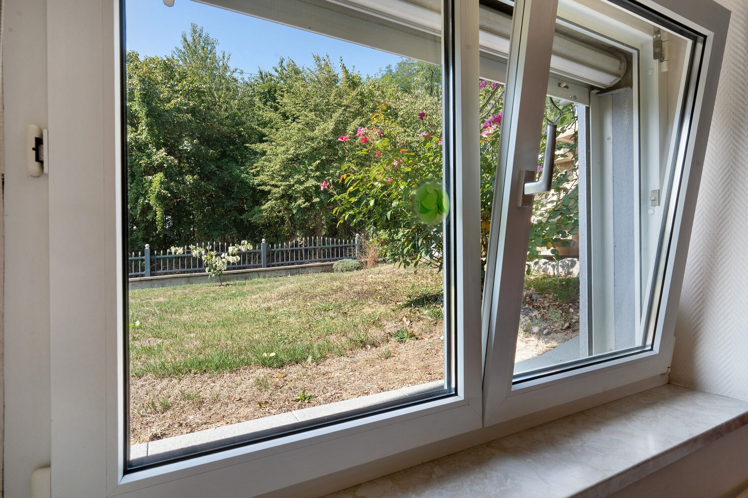 A window with a view of a garden, a metal fence, and a stone wall.