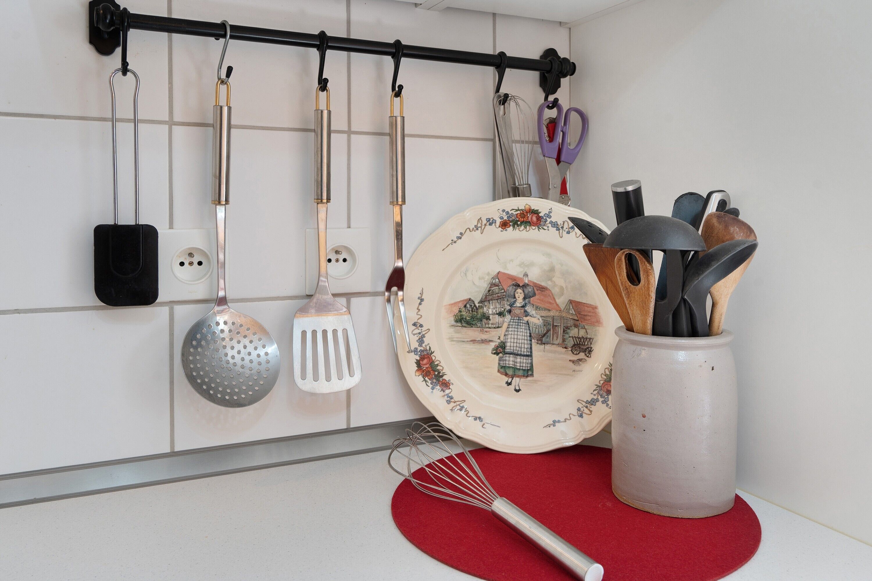 A kitchen with a utensil rack, a decorative plate, and a whisk on a red mat.