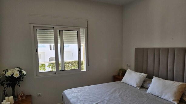 Un dormitorio con una cama, una ventana, una mesita de noche y una mesita auxiliar con un florero.
