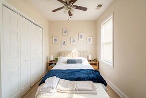 Ein Schlafzimmer mit Bett, Deckenventilator, Fenster mit Jalousien und gerahmten Bildern an der Wand.