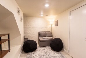 Ein kleiner, gemütlicher Raum mit weißer Ziegelwand, einem grauen Sofa, zwei schwarzen Sitzsäcken, einem Holzregal und einer Stehlampe.
