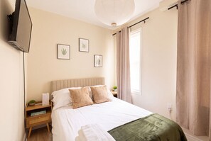 Ein Schlafzimmer mit einem Bett, einem Nachttisch, einem an der Wand befestigten Fernseher und gerahmten Pflanzenbildern.