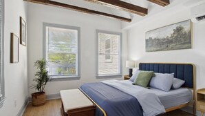 Ein Schlafzimmer mit einem Bett, einem Nachttisch, einer Lampe, einem Fenster mit Jalousien, einer Pflanze und einem Bild an der Wand.