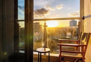 Ein Balkon mit Blick auf eine Skyline im Abendrot, ein Stuhl und ein Tisch mit einem Weinglas.