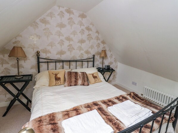 A bedroom with a bed, two nightstands, lamps, a radiator, and a patterned wallpaper. - Sciora Cottage (Crieff)