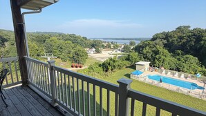 Eine Holzterrasse mit weißer Brüstung, die auf einen Swimmingpool und einen Blick auf einen See mit Bäumen und Gebäuden führt.